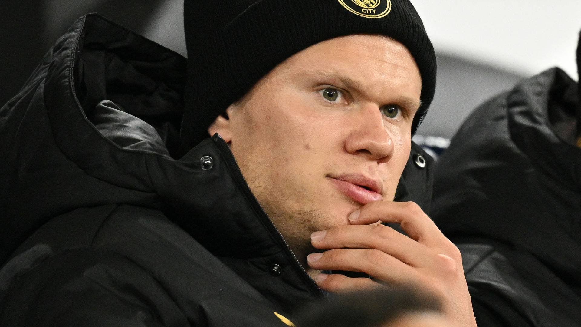 O jogador de futebol Erling Haaland, com boné preto do Manchester City e jaqueta preta, olha pensativo para a direita, com a mão direita no queixo