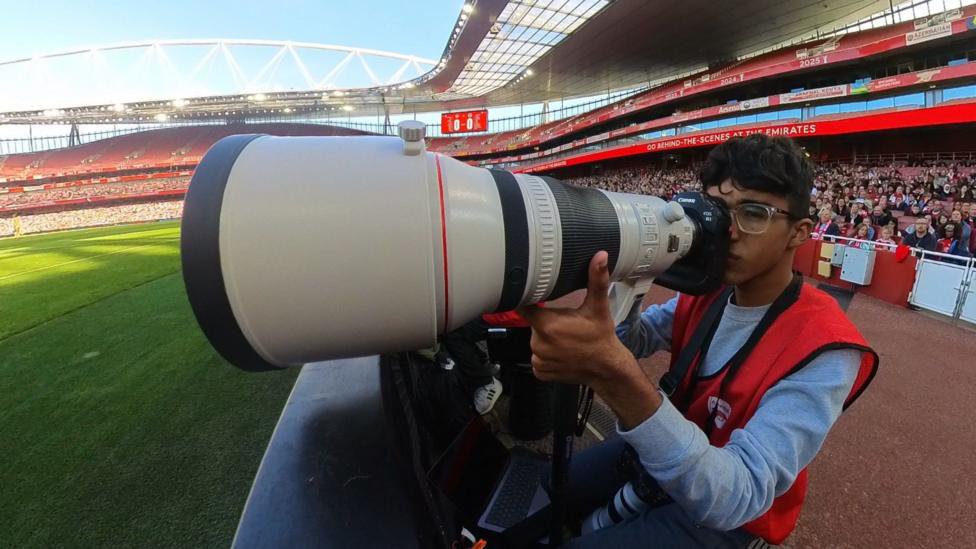 A photo of Tiego Greno, who is looking down the lens of a camera with a large grey lens attached to the front. He is sat next to the pitch at Arsenal's Emirates Stadium. He is wearing a grey jumper with a red high-visibility jacket on top.
