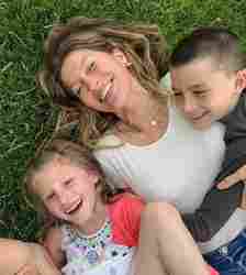 Gisele Bündchen laying in the grass with her two kids, smiling.