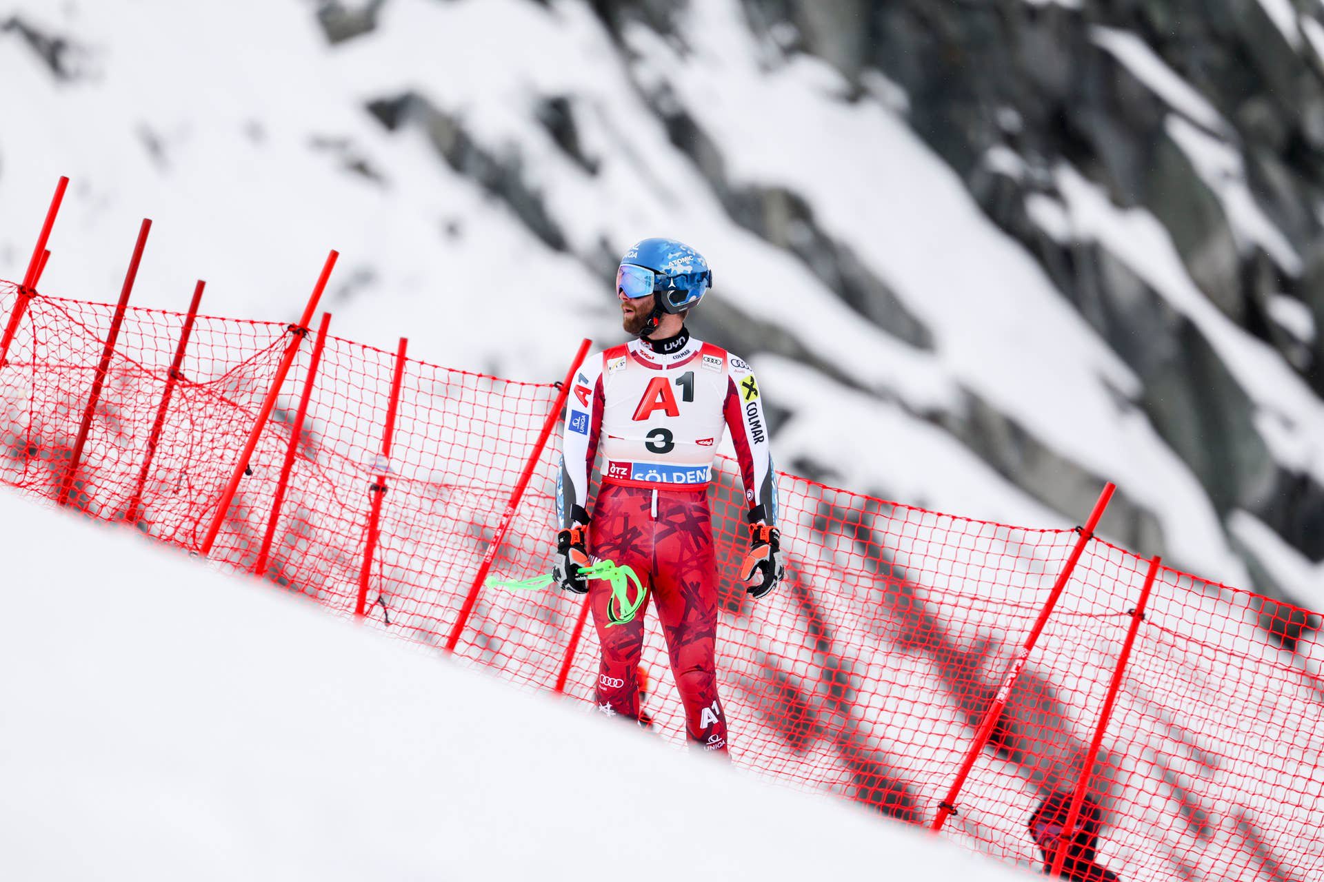 Ein Skirennläufer steht im Zielbereich vor rotem Fangzaun auf schneebedecktem Hang.