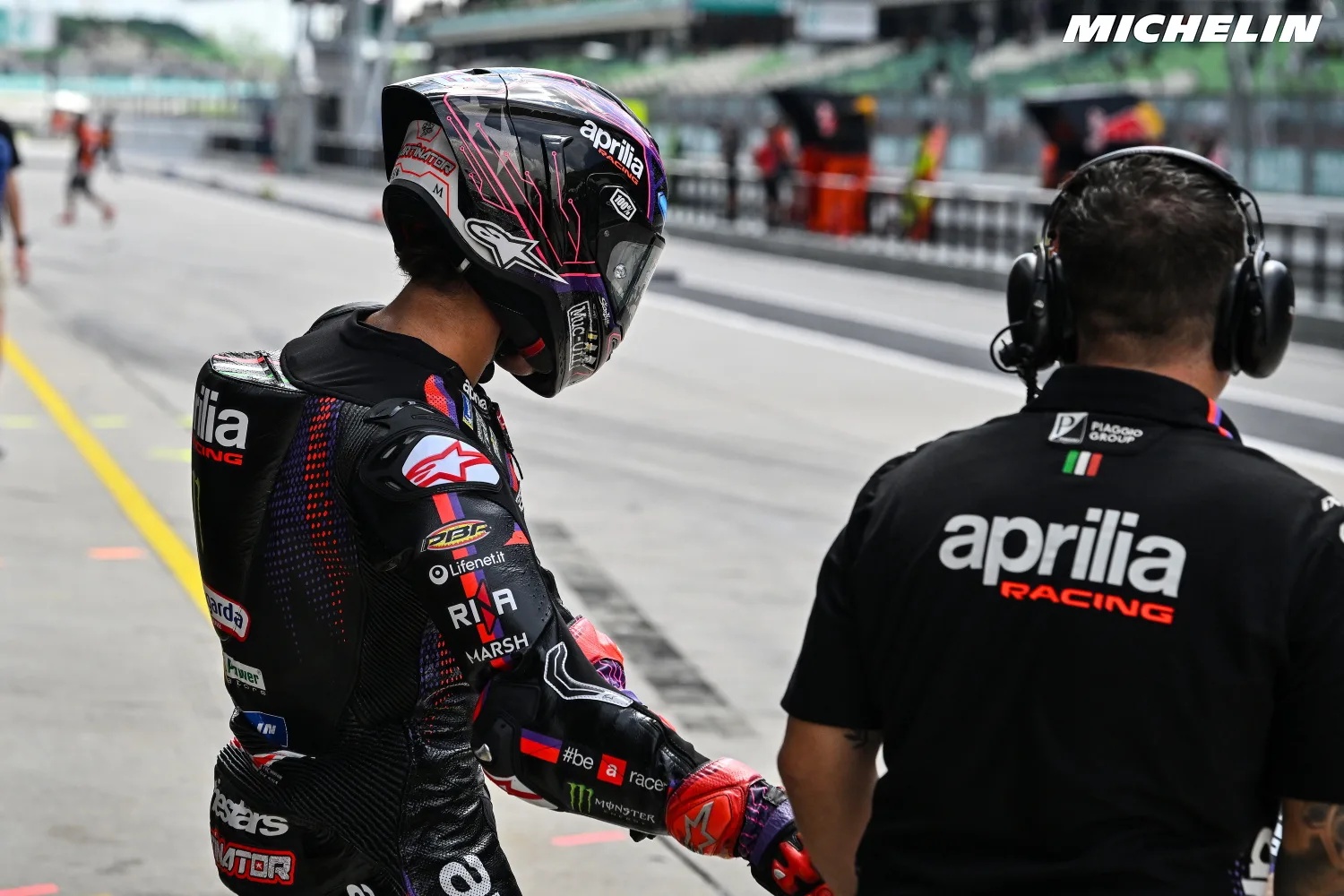 Rider in black Aprilia Racing leathers and helmet walks in the Sepang pit lane beside a crew member in an Aprilia Racing shirt, MotoGP
