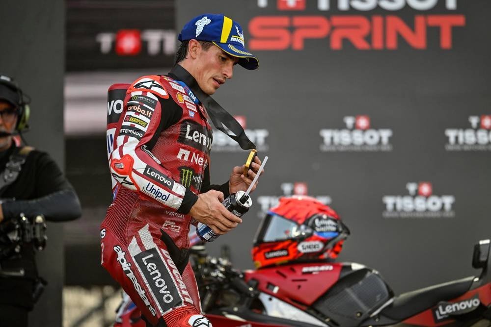 A Ducati MotoGP rider in red Lenovo-branded leathers and a cap, holding a bottle at a Tissot Sprint podium area