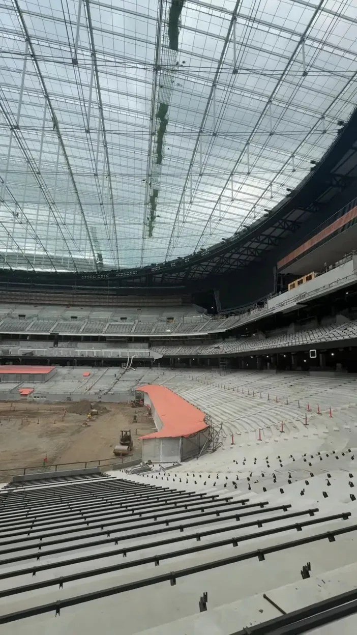 A large stadium under construction, featuring tiered seating and a sprawling glass roof.