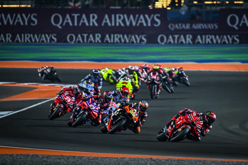 MotoGP riders racing in a pack past a Qatar Airways banner at a night race, with several red Ducati bikes leading