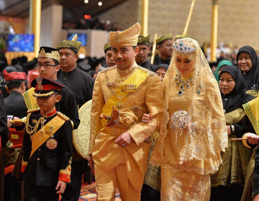 Royal couple in traditional golden attire walk with attendants in a ceremonial setting.