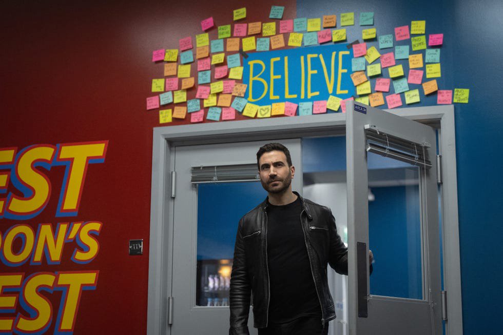 A man in a black leather jacket stands at a door with a wall of colorful sticky notes above labeled "BELIEVE.