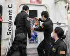 Four NASA personnel in black jumpsuits and face masks are seen placing a mission patch on the door of a spacecraft module labeled "Segment C." An American flag is visible above the door.