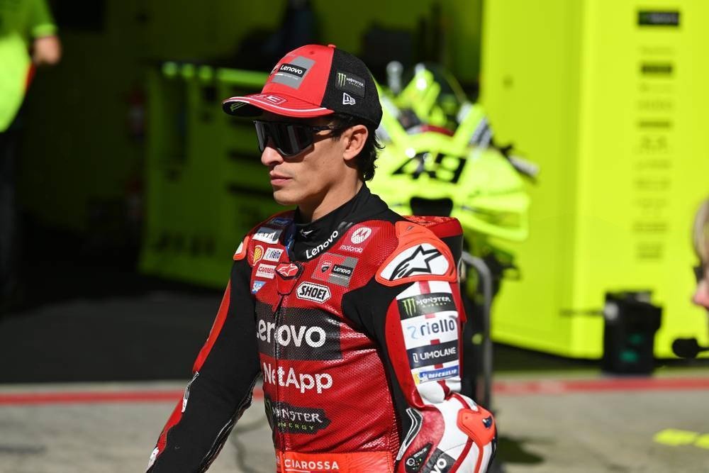 A Ducati MotoGP rider in red and black leathers and cap in the paddock, with a yellow pit garage behind him