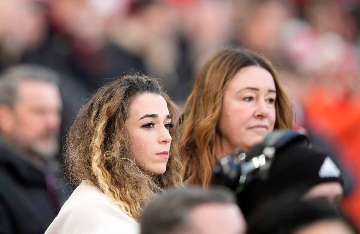 Two women with long hair are looking to the left, seemingly focused on something in the distance. They are situated in a crowded public setting, possibly at a sporting event, as indicated by the presence of spectators behind them. The surrounding audience members are partially visible, creating a sense of depth and activity.