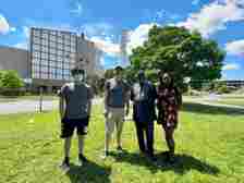 Four people stand on grass outside a grey concrete building on a sunny day.
