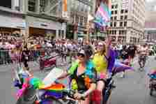 Two women on a motorcycle on the parade route wearing colorful clothing and waving a trans flag
