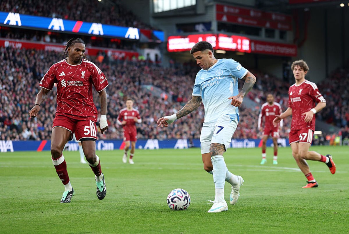 Two soccer players in red and light blue jerseys compete for the ball on a green field during a match.
