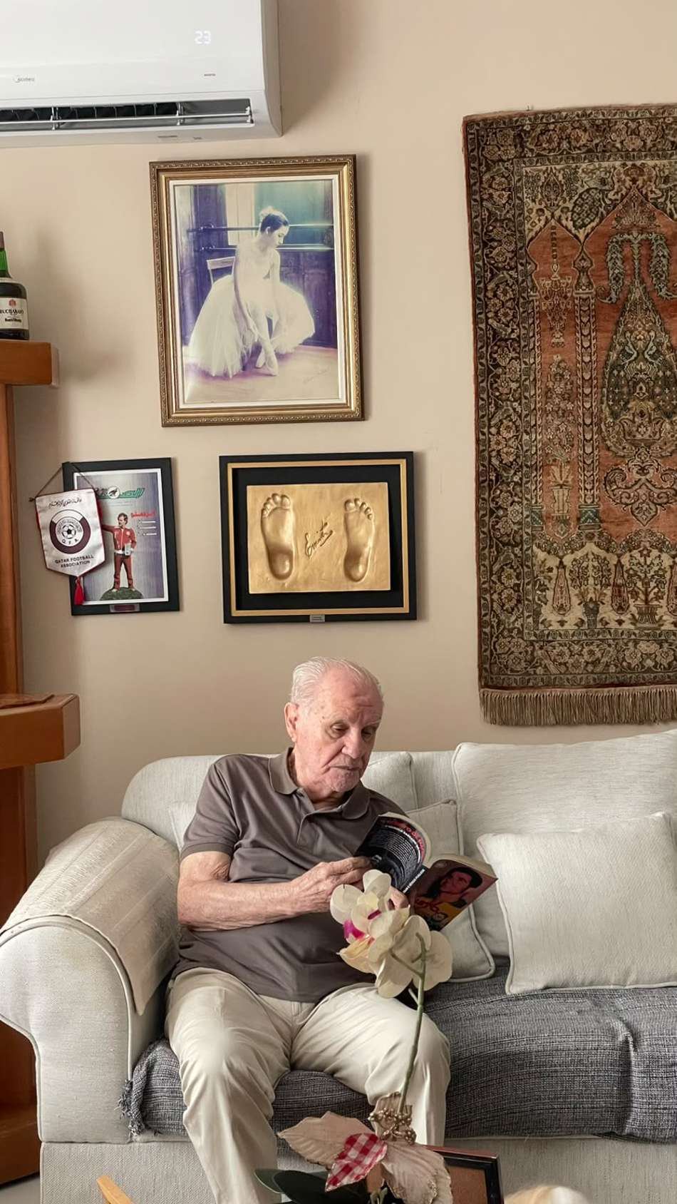 Evaristo Macedo lendo livro no sofá, em sala com quadros na parede e tapeçaria ao fundo.