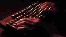 Person's hands typing on a black keyboard with red light in dark room.