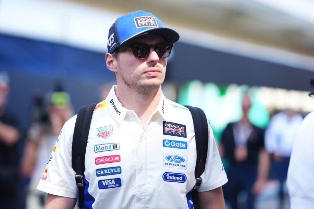 A driver in a white Oracle Red Bull Racing shirt and blue cap with sunglasses, standing in the F1 paddock at COTA media day.