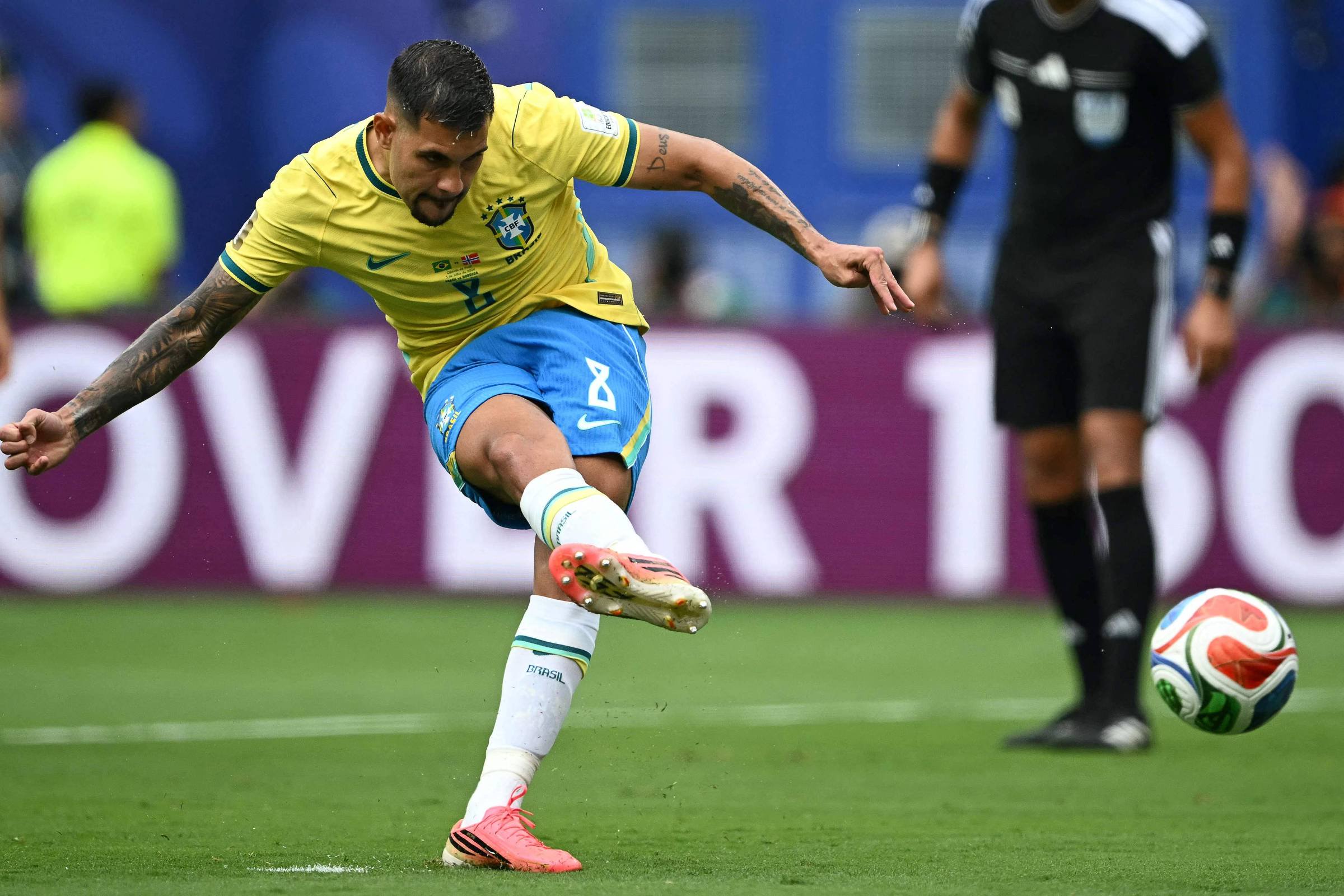 Jogador da seleção brasileira com uniforme amarelo e azul chuta a bola com o pé direito em campo de futebol. Ele é Bruno Guimarães. Árbitro vestido de preto observa a jogada ao fundo.