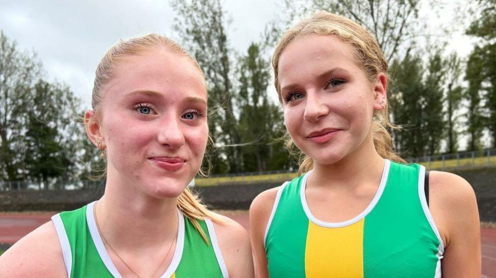 Two blonde teenage girls standing next to each other on a race track wearing green and yellow sports gear.