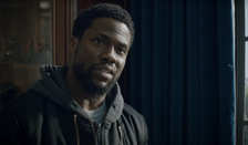 Kevin Hart in a casual jacket and hoodie, looking serious, in an indoor setting with curtains in the background