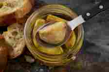 A jar of roasted garlic cloves in oil with a spoon lifting one clove, surrounded by slices of toasted bread