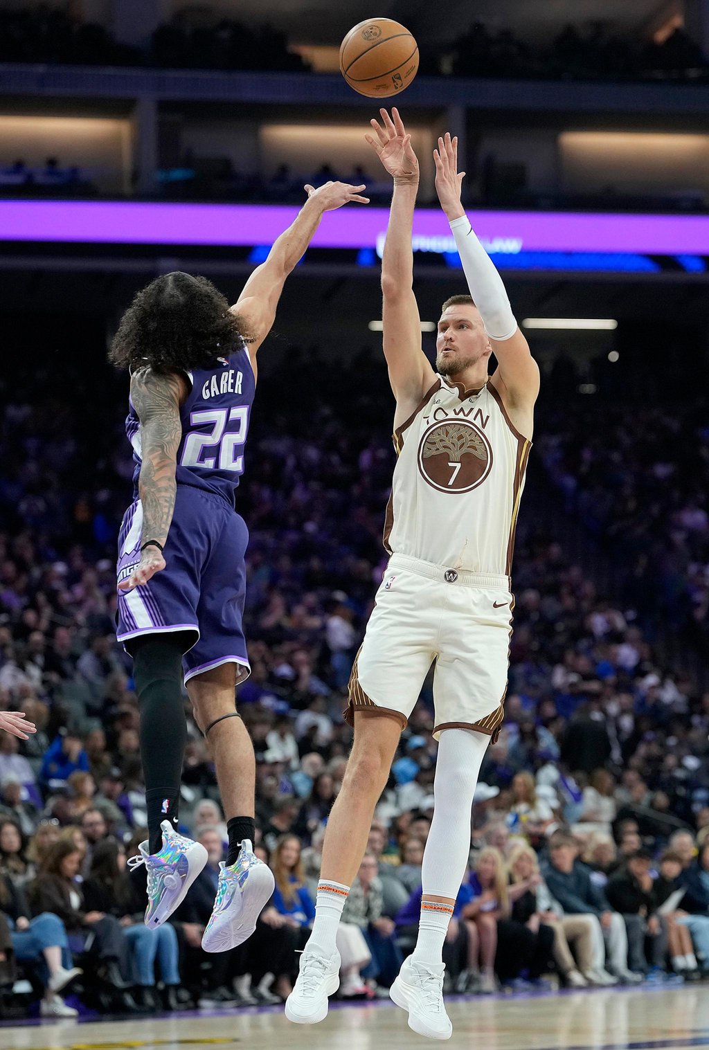 A basketball player in a white and brown uniform shoots over a defending player in a purple uniform.