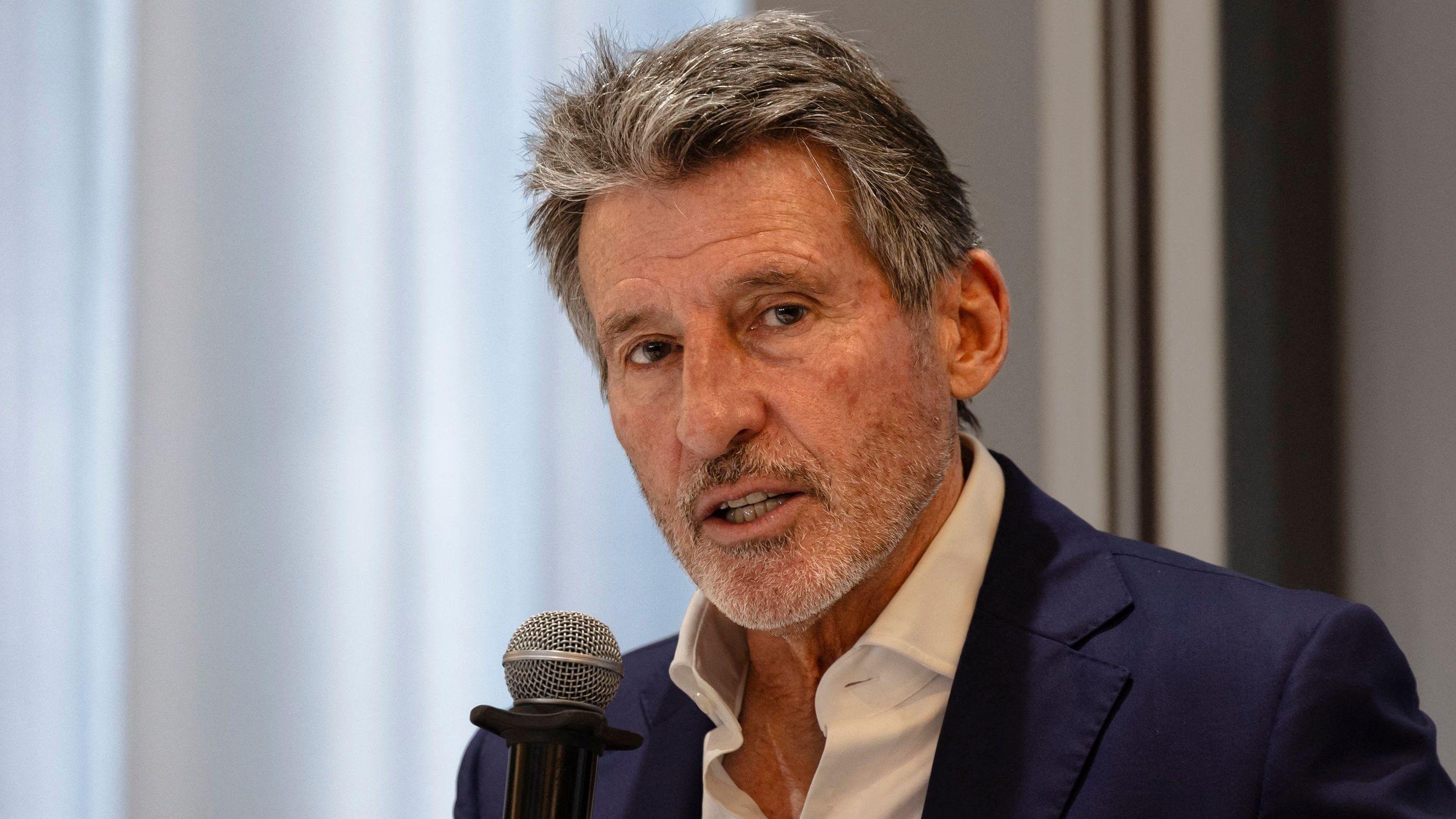 Sebastian Coe, visible from shoulders up wearing a white open-necked shirt and a navy blue suit jacket, holds a microphone up to his mouth as he speaks at a news conference. A wall and a window can be seen out of focus behind him