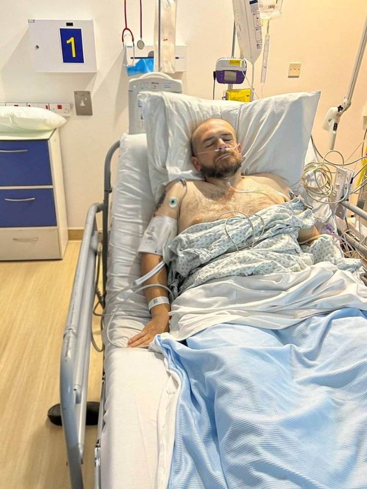 An individual is lying on a hospital bed with medical equipment and accessories around them, indicating a medical setting.
