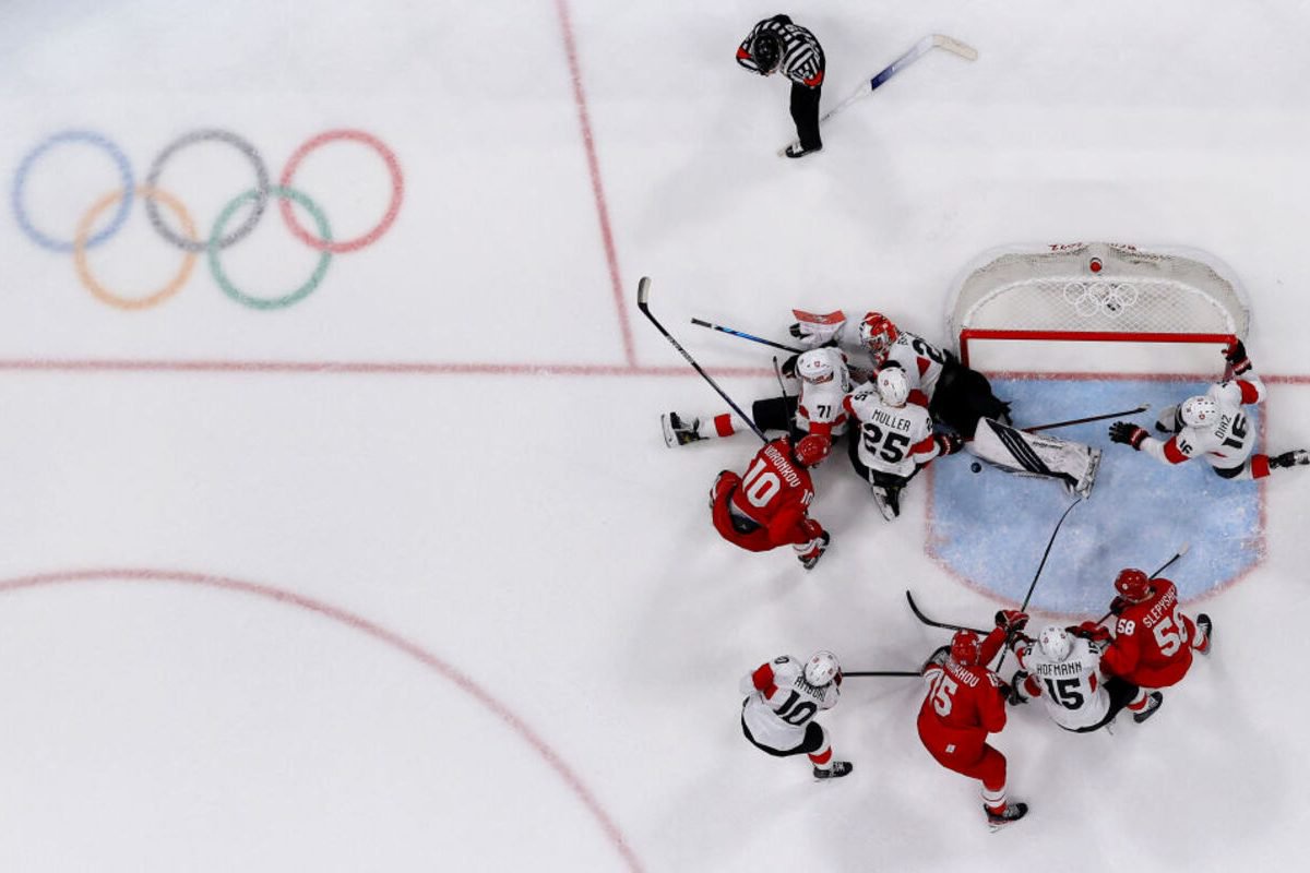 Olympic hockey players in red and white jerseys scramble around the goal, with a referee watching nearby.