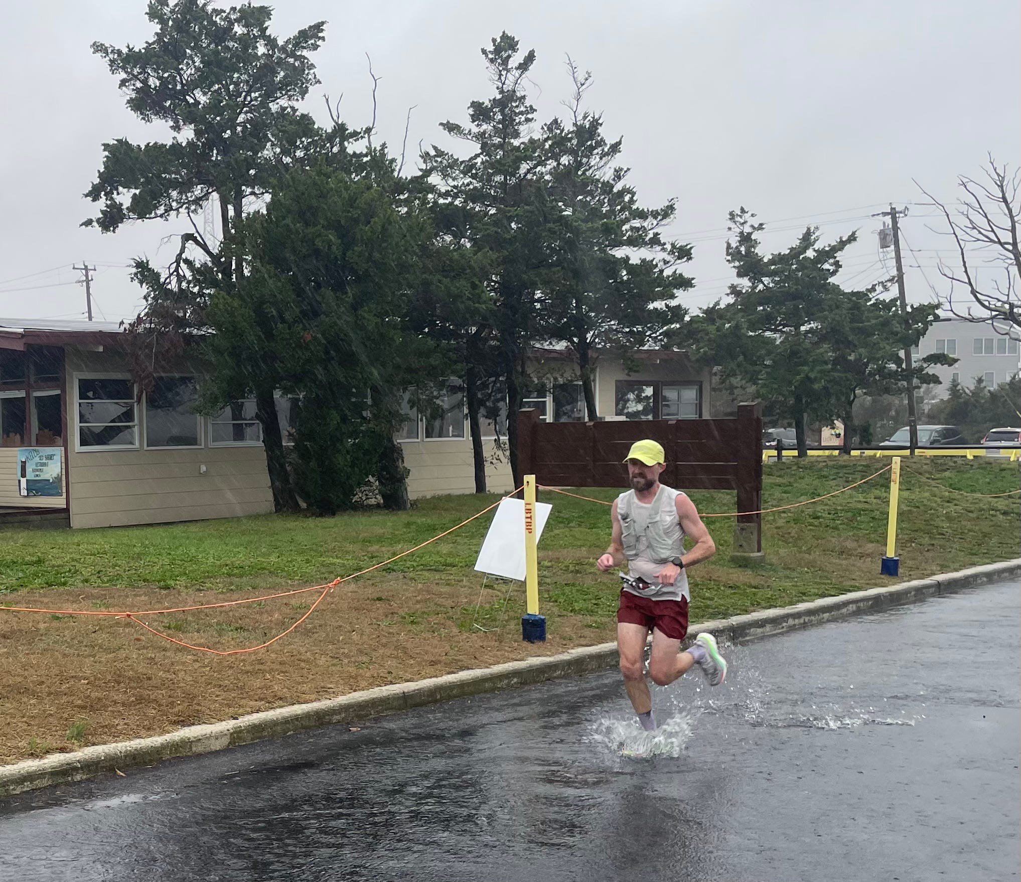 A runner splashes through a puddle during a rainy day race near a building and trees.