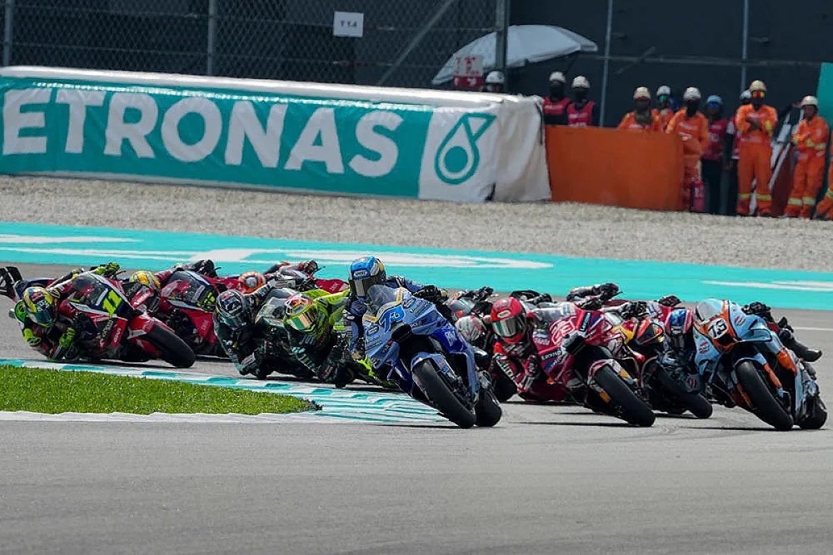 MotoGP riders on track at Sepang, with a Petronas-branded barrier and a rider in blue and white racing ahead of the pack