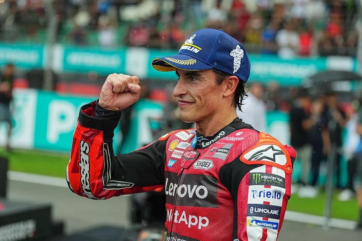 A MotoGP rider in red and black Aprilia-style leathers with Lenovo and Alpinestars logos raises a fist and smiles in the Sepang paddock area
