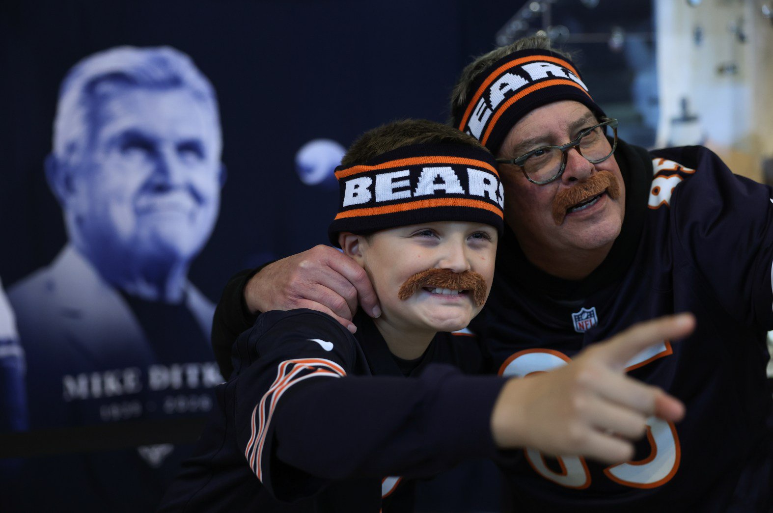 Kieran Checketts, 12, and his grandfather, Tom Borich, wear Mike Ditka-like fake mustaches while taking pictures at a display remembering the former Bears head coach and player on Oct. 22, 2026, before the  Bears host the Patriots at Soldier Field. It was the first home game since Ditka’s death and a Bears homage included a stadium-wide Ditka mustache giveaway and a knit headband modeled on Ditka’s iconic sweater vest. (John J. Kim/Chicago Tribune)