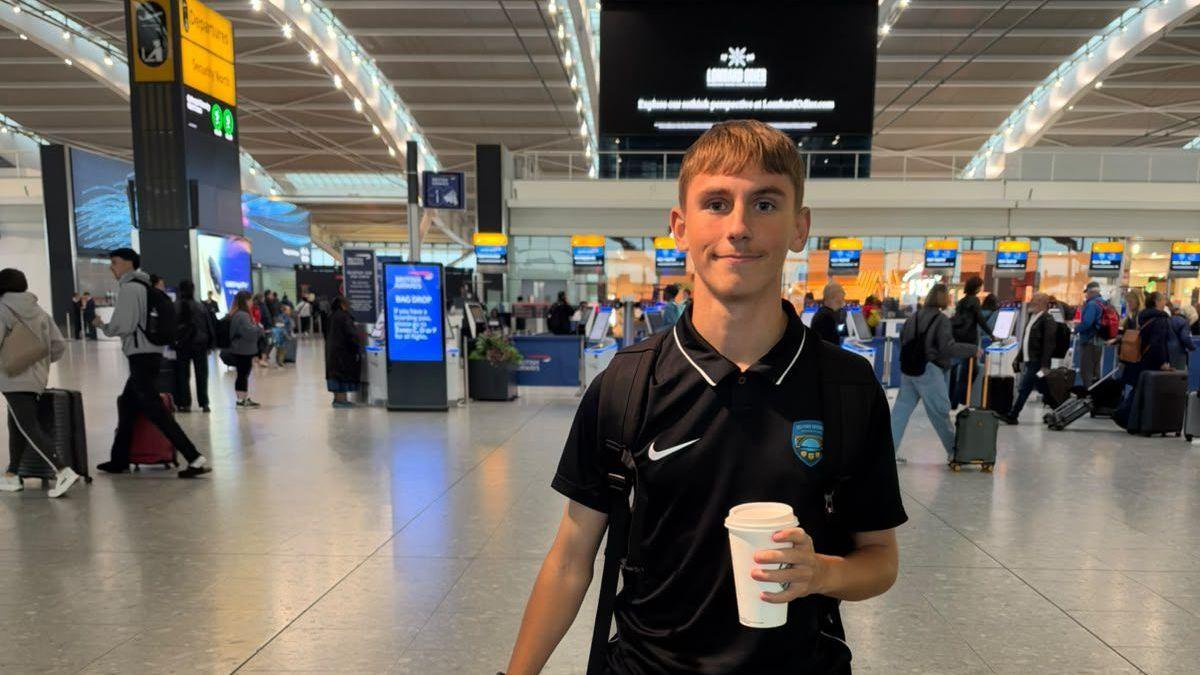 A young man with short fair hair and a black T-shirt holding a white coffee cup in a grey airport departures area