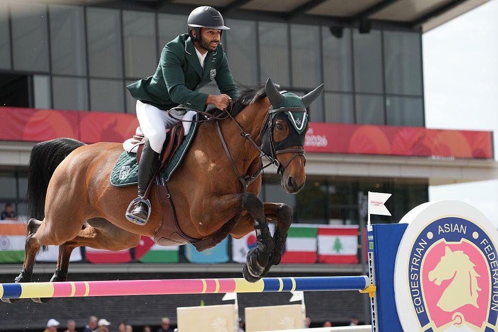 Saad Abdullah S Alajmi of Saudi Arabia and horse clear a jump during an equestrian event. Alajmi wears a dark green jacket, white breeches and a helmet, while the horse is fitted with protective leg wraps and a green ear bonnet.