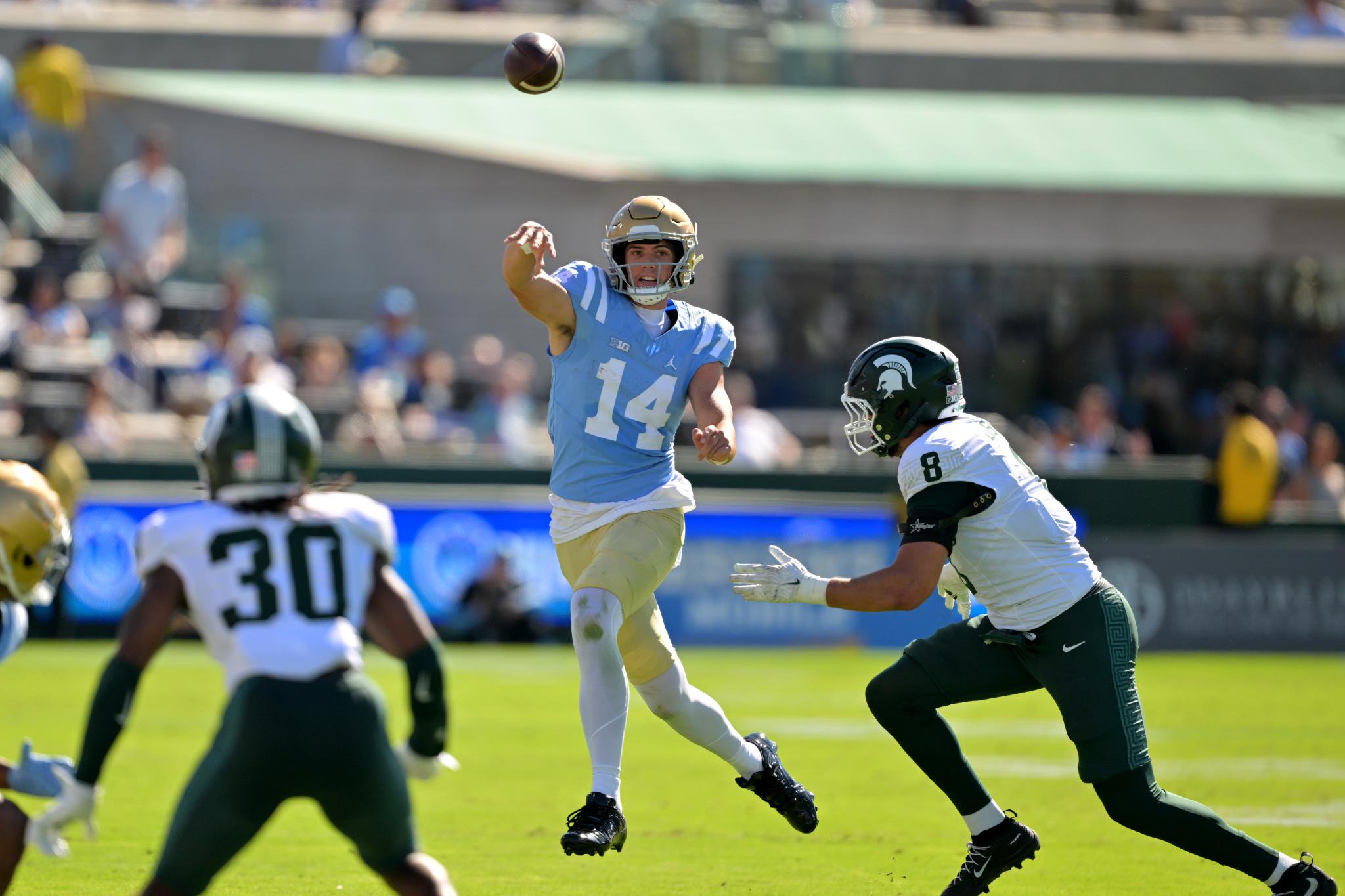 A UCLA quarterback throws a pass under pressure from Michigan State defenders during a game.