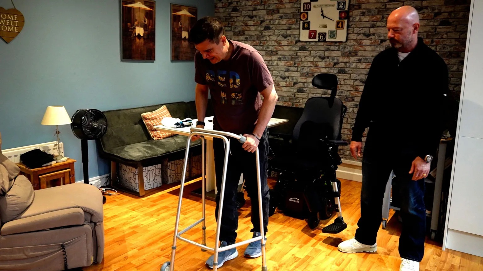 BBC Keith Dungait using a walking frame. He has short dark hair and is wearing a brown t-shirt and dark trousers. There is a wheelchair behind him. To his left is Alan Shearer, wearing a dark fleece, dark trousers and white trainers. The pair are in a living room setting, with a sofa and armchair and and exposed brick wall behind them.