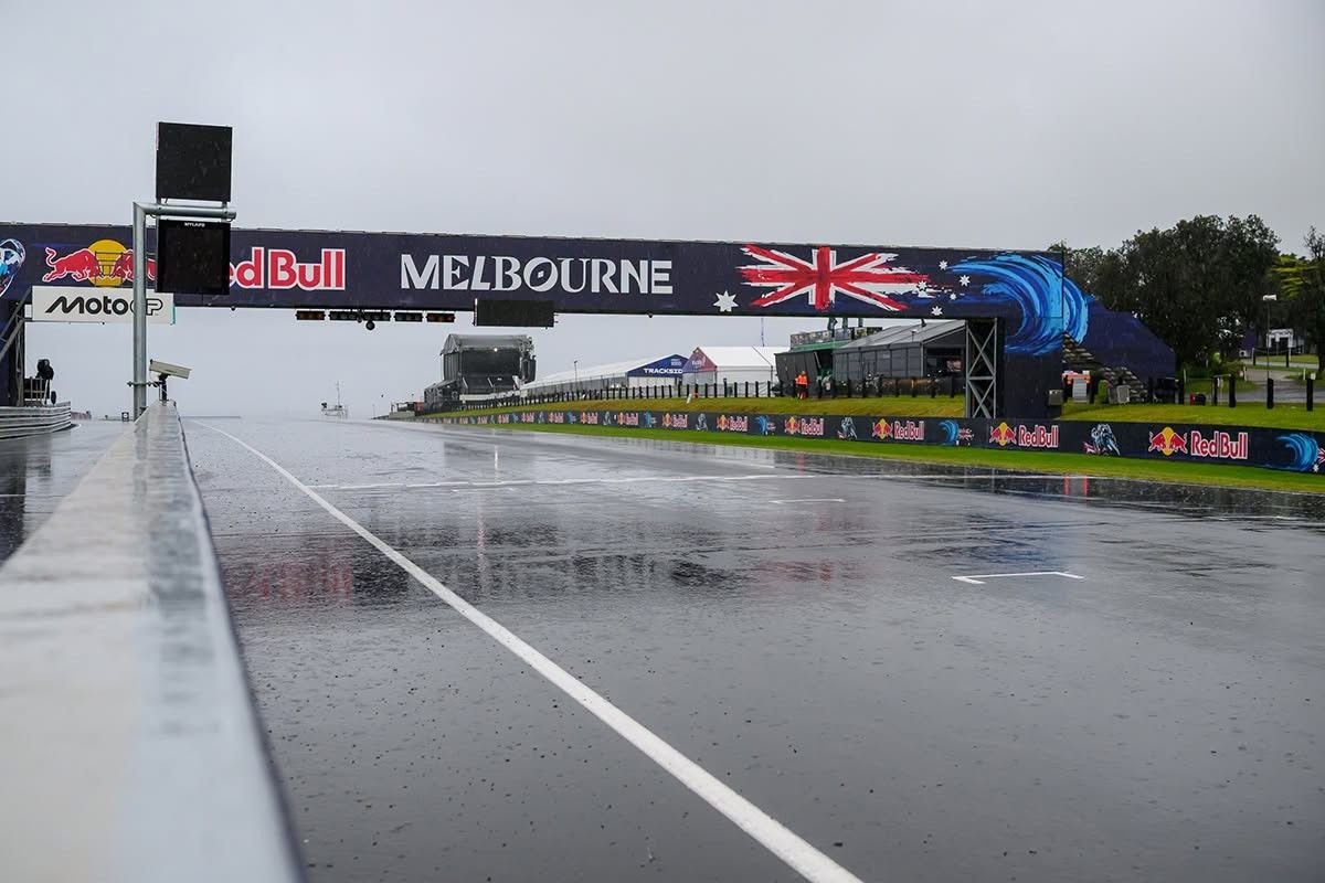Wet MotoGP track at Phillip Island under a "Red Bull Melbourne" gantry, with a Moto GP sign and empty grid, rain pooling on the asphalt