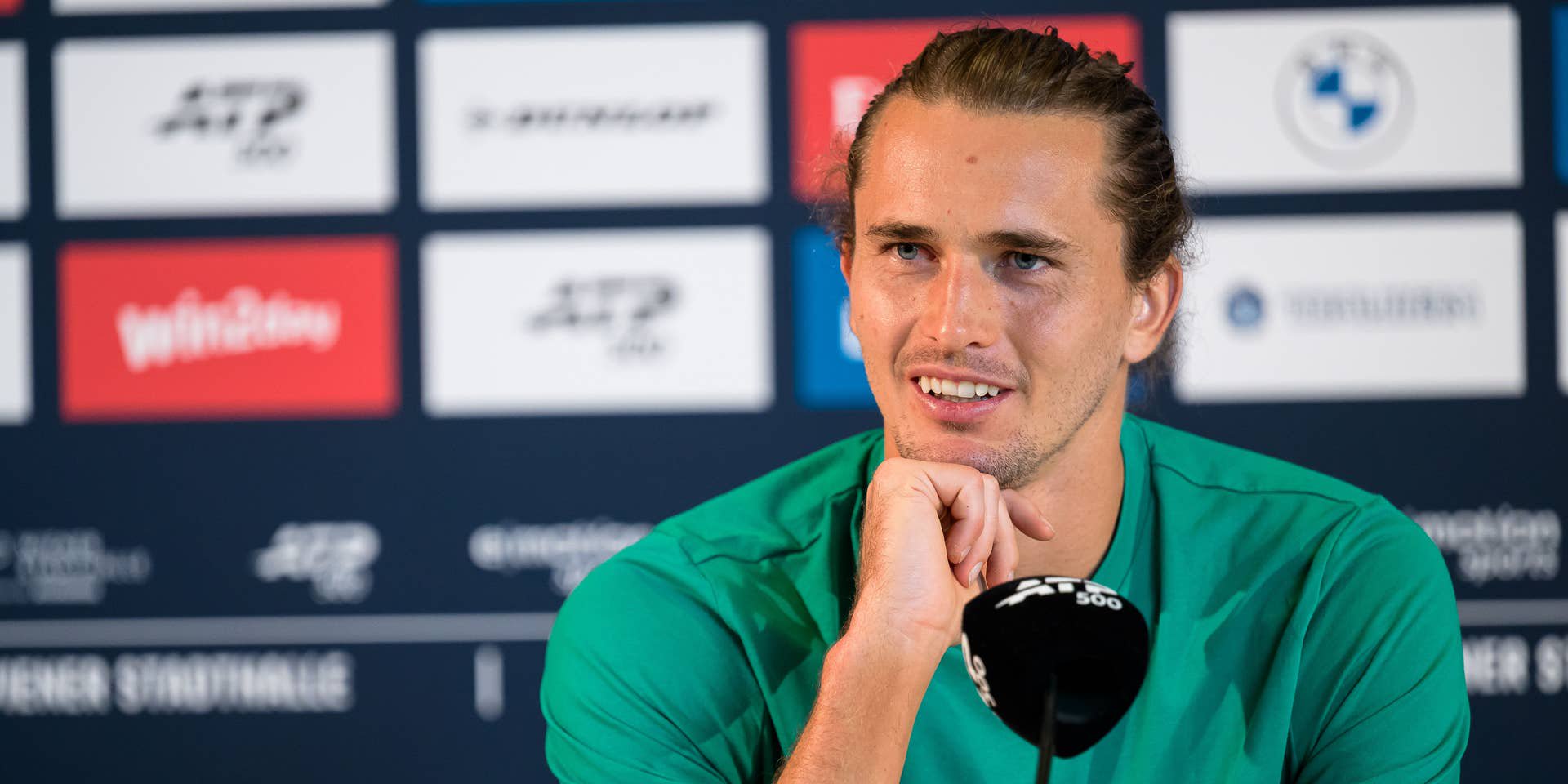 Ein Mann im grünen Shirt spricht lächelnd bei einer Pressekonferenz vor einem Mikrofon.