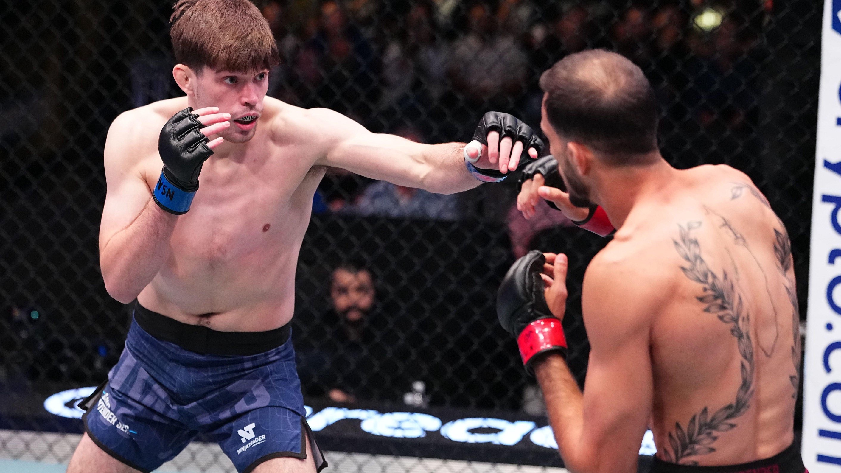 Two mixed martial artists face each other inside a cage. The competitor on the left, wearing blue shorts and black gloves, is punching towards his opponent who is wearing red shorts and black gloves. The arena crowd is blurred in the background.