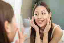 Fresh healthy skin, beautiful smile of asian young woman, girl looking at mirror, applying moisturizer on her face, putting cream treatment before makeup cosmetic routine at home. Facial Beauty.