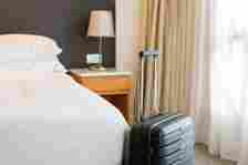 A neatly made hotel bed with a side table and lamp next to it. A black suitcase is placed upright near the bed. 
