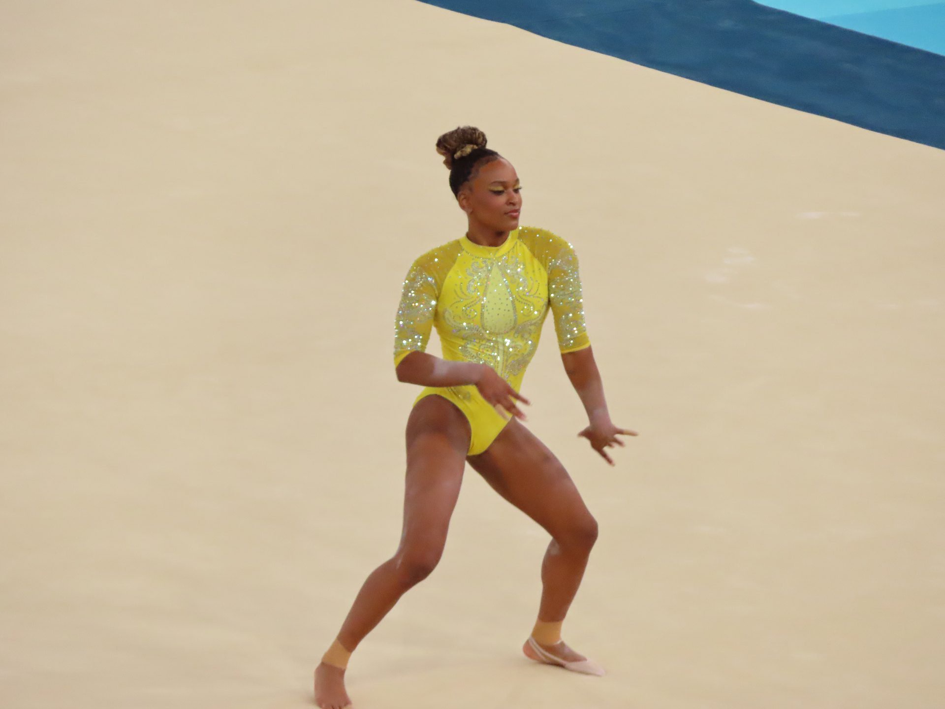 Ginasta brasileira Rebeca Andrade, vestindo collant amarelo brilhante e com o cabelo preso em coque, realiza movimento coreográfico sobre o tablado de ginástica artística nos Jogos Olímpicos de Paris 2024