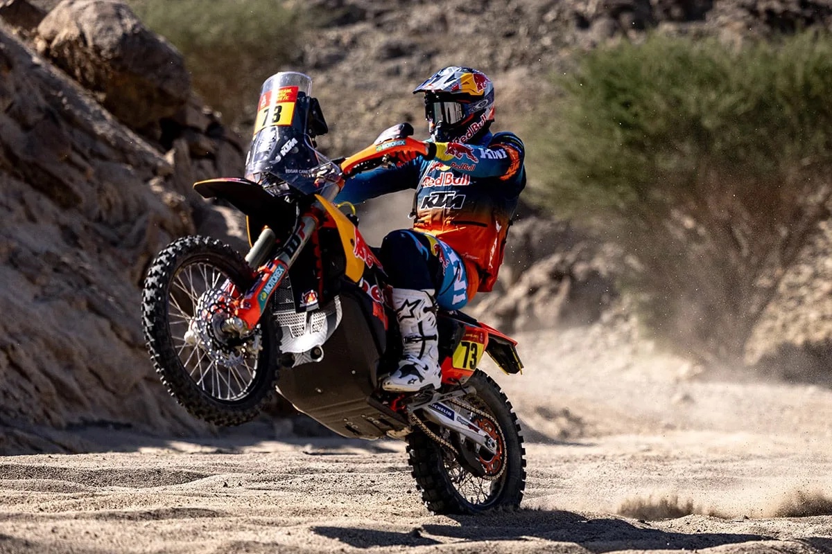 Rider in Red Bull KTM orange and blue leathers, race number 13, wheelie on a KTM rally bike through a desert wadi, Dakar Rally