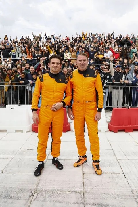 Two race car drivers in bright orange racing suits stand in front of a cheering crowd behind a barrier.