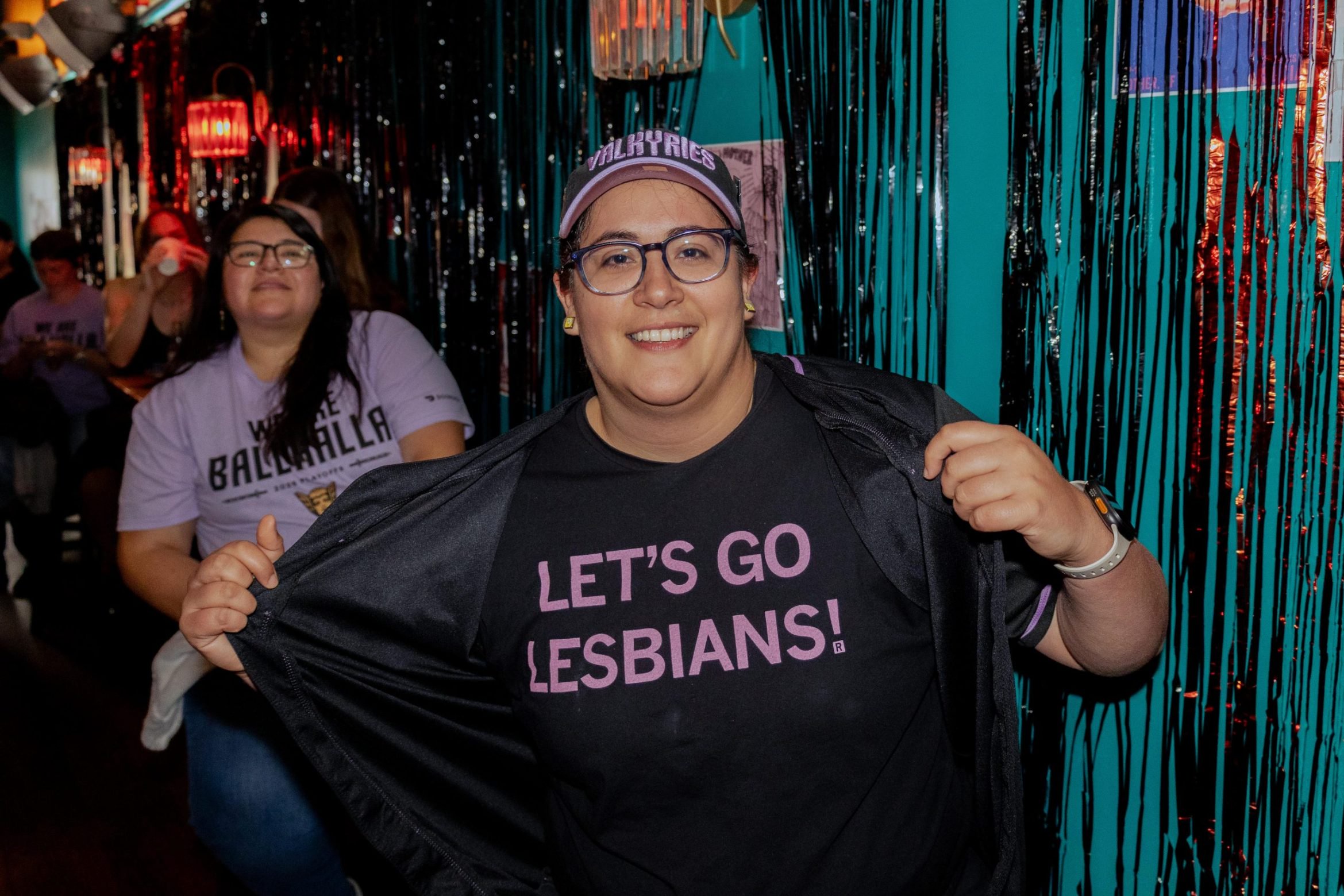 A person wearing glasses and a cap holds open their jacket to reveal a black t-shirt that says “LET’S GO LESBIANS!” in pink text.