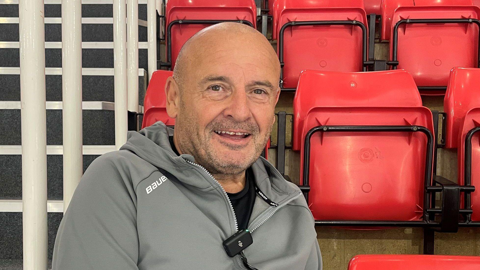 Image of a man wearing a grey top, sitting in a sports stadium and smiling at the camera