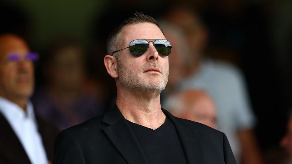 Peterborough United chairman Darragh MacAnthony looking on from the stands wearing a black blazer, a black shirt, and black sunglasses