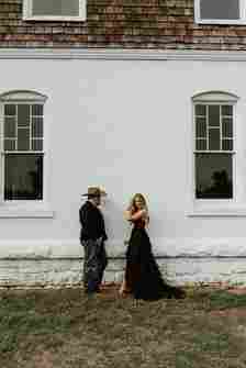 A man wearing a cowboy hat, black jacket, and jeans stands facing a woman in a long, flowing black dress. They are standing in front of a white brick building with two large windows. Both figures are looking at each other, with the woman holding her hand near her face.