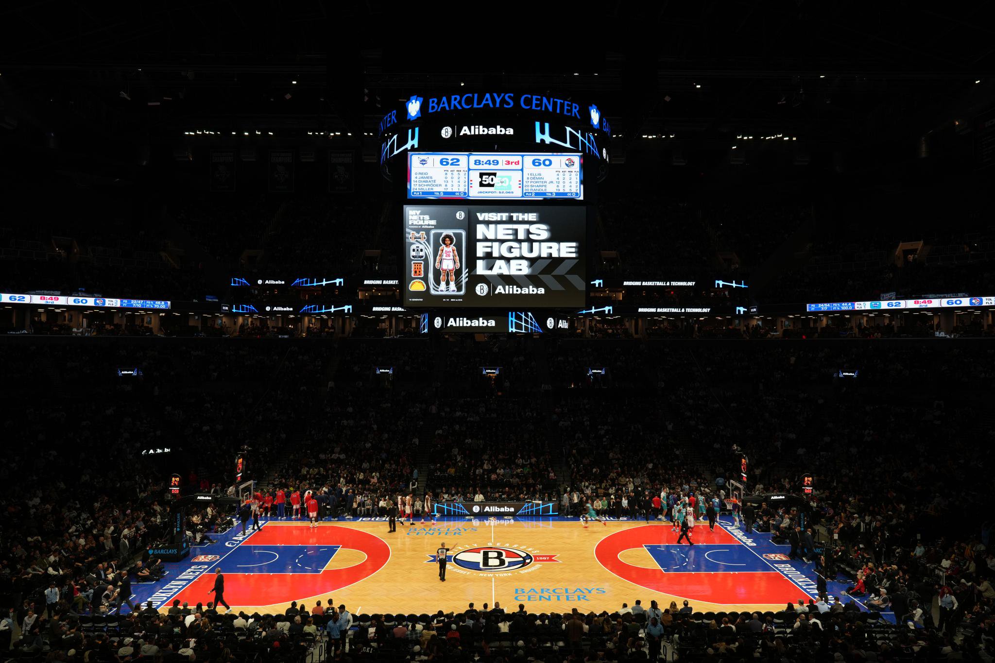 Wide shot of a basketball game at Barclays Center, showing the glowing court, packed stands, and the large central scoreboard displaying game stats and promotions.