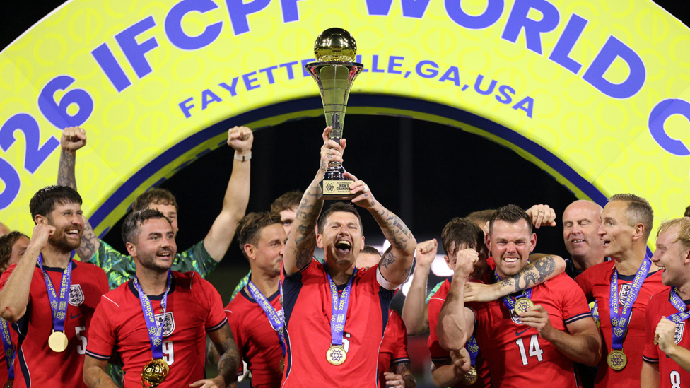 A team photo of England, in red shirts, lifting the Cerebral Palsy World Cup Trophy aloft.