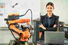 A woman in business attire stands confidently beside industrial robotic machinery in a tech workspace